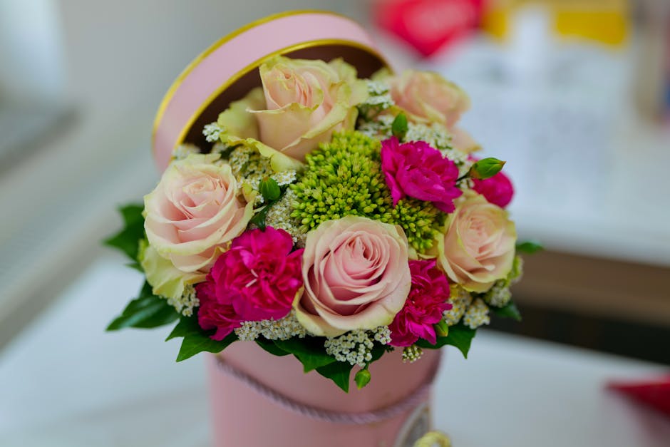 A floral bouquet arranged in a round, pale pink cylindrical box with a pink rim, featuring soft blush pink roses, vibrant hot pink carnations, green hypericum berries, and delicate white baby's breath