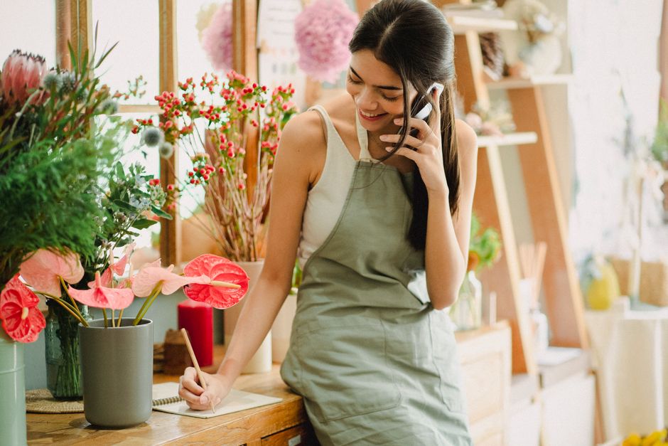 A smiling young woman with long dark hair, wearing a beige apron over a white tank top, is standing behind the counter of a florist shop. She is holding a mobile phone to her ear with her right hand w