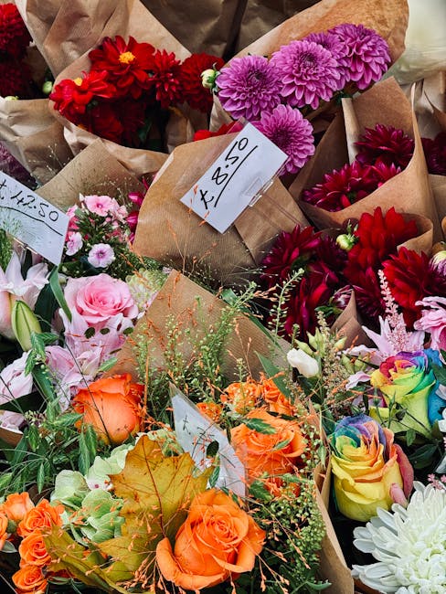 A vibrant display of fresh flowers, including bright pink chrysanthemums, deep red dahlias, peach roses, rainbow-hued roses, and delicate white blooms, all arranged in paper wrapping and placed on a t