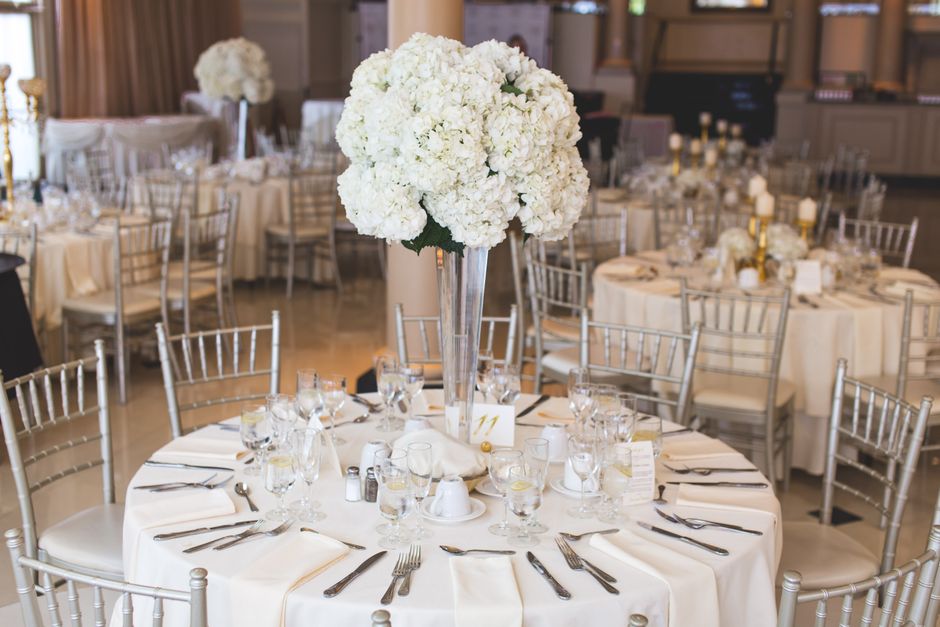 A large round wedding reception table decorated with a tall, elegant glass vase holding a bouquet of densely packed white hydrangeas, creating a lush, rounded floral centerpiece. The table is draped w