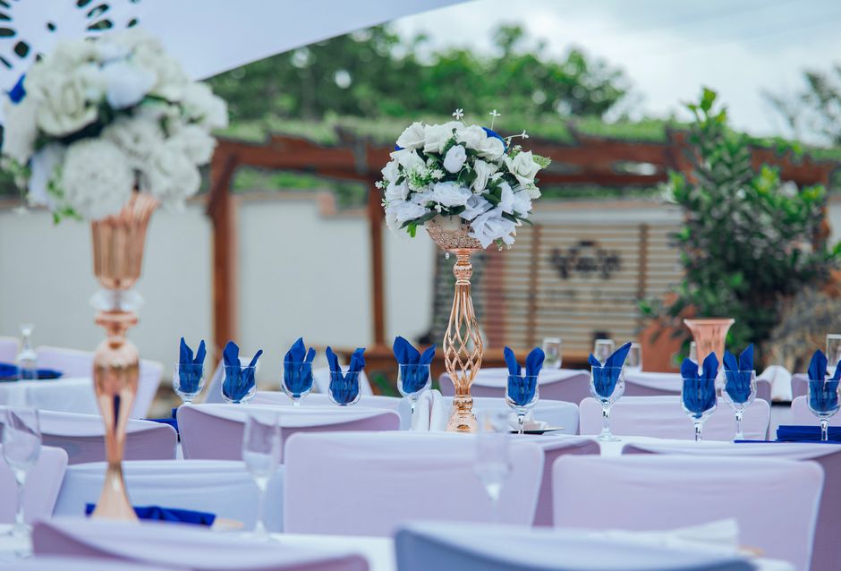 An outdoor wedding reception setup featuring tall, elegant floral arrangements with predominantly white blooms, including roses and hydrangeas, placed in ornate, rose gold vases atop white-covered tab