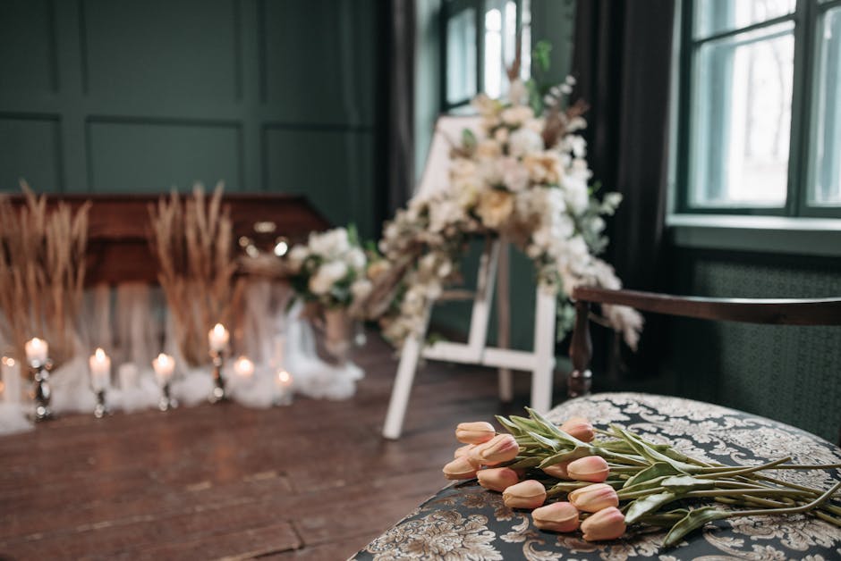A somber floral arrangement featuring soft pink tulips laid on an ornate patterned cushion in the foreground, with a blurred background of a funeral setting that includes a large display of white flow