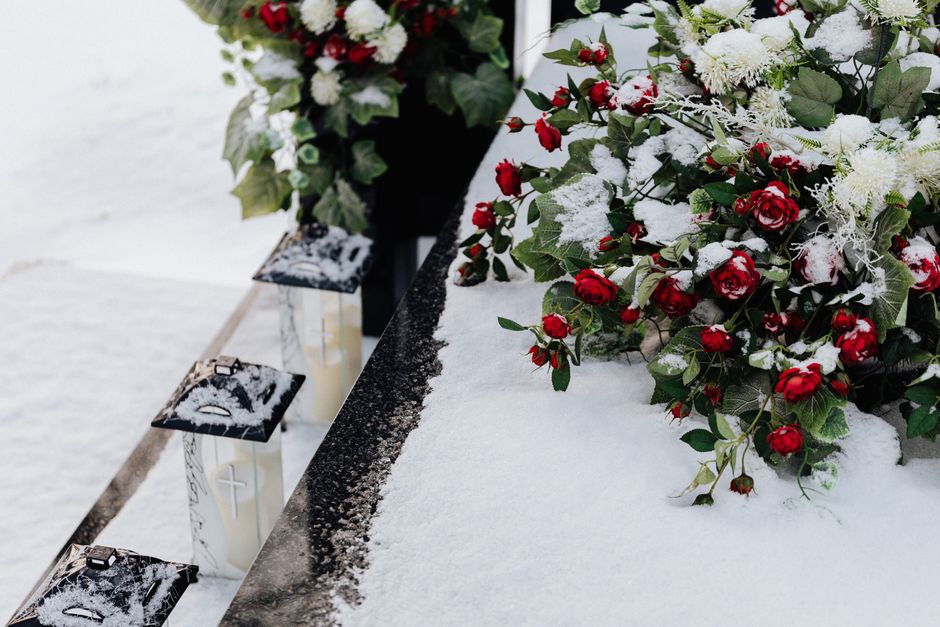 A floral arrangement featuring vibrant red roses and white flowers, partially dusted with snow, resting on a snow-covered surface. The bouquet is wrapped in a decorative green foliage, with some flowe