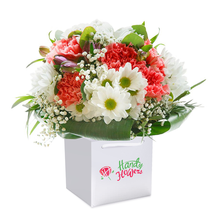 A vividly detailed bouquet sits poised and upright in a clean-lined, square white gift bag, photographed against a spotless white background that draws the eye to its nuanced palette and floral craftsmanship. Bursting with ruffled coral, peach, and red carnations, their layered petals create depth and tactile richness, reminiscent of the textures found in Tolworth's Friday flower stalls. Bright white daisies with sunny, lime-green centers nestle in the arrangement, bringing uplifting contrast and a whisper of classic charm. Wisps of baby's breath thread delicately throughout, their tiny stars adding lightness and serene movement. Unopened buds-touched with pale purple and pink-appear along the edges as gentle accents, suggesting both anticipation and seasonality. Enveloping this profusion, deep green, bold foliage forms an elegant natural border, while broad leaves wrap securely around the base, echoing the bouquet's sturdy, full form. The bag's white ribbon handles stand ready for carrying along Ewell Road. Each flower and leaf tells a story of careful selection and placement; together they form a lush silhouette, balanced and inviting. Soft, diffused daylight washes across the petals and greens, hinting at early mornings in local gardens. This arrangement makes for a cherished birthday gift, an anniversary surprise, or a thoughtful gesture delivered right to a neighbour's door in Tolworth. Every aspect speaks to the skill and passion of local florists, celebrating both nature's bounty and the artistry in every choice.