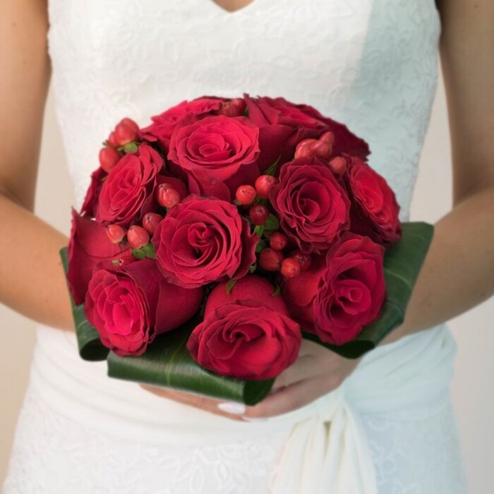 In this evocative Tolworth bridal portrait, a close-up reveals a bride's waist and arms, her posture both serene and purposeful. Her pristine white wedding dress, adorned with a lace bodice in delicate floral motifs and a gentle V-neck, frames a bouquet that instantly commands the senses. The arrangement, densely packed and perfectly symmetrical, boasts a profusion of deep scarlet red roses-some petals elegantly furled, others open in soft spirals, each glistening faintly under gentle daylight. Interwoven among the blooms are bright red clusters of hypericum berries, their polished surface lending a playful, jewel-like dimension and a touch of local celebration. At the bouquet's base, glossy dark green foliage provides sculptural support and a cool, refreshing contrast to the roses' rich warmth. The bride's hands, thoughtfully cupped beneath the stems, reflect skilled local floristry and the meaningful ritual of Tolworth weddings. The blurred, softly lit background falls away, allowing every velvety petal and textured berry to take centre stage-a tribute to enduring romance, abundance, and elegant craftsmanship. This image speaks not only to timeless wedding tradition but to the unique flair and freshness of weddings along Tolworth's leafy avenues, perfect for those seeking inspiration for their special day in this vibrant community.