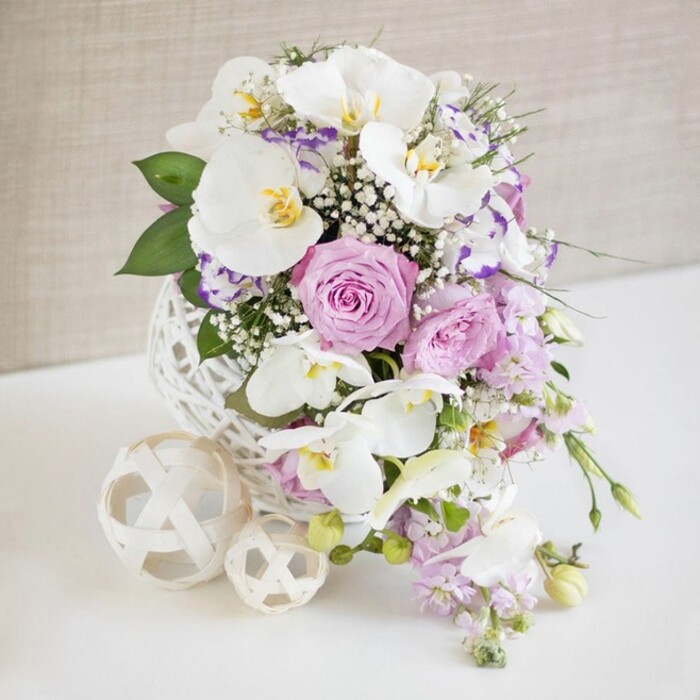 A richly detailed floral arrangement sits centered on a white surface, as if awaiting placement in a Tolworth wedding venue or a beautifully laid family celebration table. The composition is rooted in a striking white spherical vase whose open, woven design feels both modern and handcrafted, its interlacing strands forming a delicate lattice like a ball of carefully looped wicker. Rising from this sculptural base is an abundant bouquet in shades of white, soft pink, and lavender, with cooler purple tones that lend sophistication. The eye is immediately drawn to several large white Phalaenopsis orchids, their smooth, broad petals fanning out gracefully, while hints of yellow and golden tones glow at their centers, adding a whisper of warmth. Around these orchids, velvety roses in tender blush pink and muted lilac swirl into full bloom, each flower layered with intricate petals that suggest romance and careful craftsmanship. Interspersed among the roses are smaller, ruffled blossoms edged delicately in purple or fuchsia, likely Lisianthus or Eustoma, which add fine detail and gently deepen the colour story. Airy sprays of baby's breath weave throughout the bouquet, their tiny white florets forming a soft veil that lightens and expands the arrangement visually. From the lower edge of the design, slender sprigs of light purple stock or similar trailing flowers extend downward, imparting a touch of movement, as though the flowers were gently flowing toward the table. Pockets of fresh green leaves peek through, bringing a natural contrast to the pastel hues. To one side, two decorative woven balls in a pale cream shade, one larger than the other, rest on the tabletop, mirroring the vase's open lattice and providing a sense of harmony. Set against a softly textured beige backdrop and illuminated with clear, even light, this design by Flowers Tolworth feels ideal for same-day delivery to local receptions, small gatherings, or elegant home dining tables across the Tolworth area.