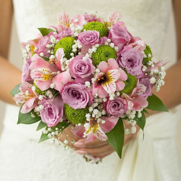 Bringing the refined artistry of Flowers Tolworth to life, this close-up showcases an exquisite, hand-tied bridal bouquet gently clasped against the delicate detailing of an ivory wedding gown. Lush, rounded, and impeccably balanced, the bouquet is centered on a medley of soft lavender and blush pink roses, some adorned with tiny droplets that sparkle softly in the bridal suite's filtered light. Interwoven throughout are flawless lime green button chrysanthemums, adding a playful and fresh counterpoint to the romantic roses. Pink alstroemeria lilies infuse the arrangement with an exotic charm-each trumpet-shaped bloom marked by intricate maroon and gold streaks. Wisps of dainty white baby's breath create a cloudlike texture, filling the bouquet with airy volume and lightness. A few deep green leaves peek through at the edges, adding natural structure and definition. The bride's fingers, adorned with a simple ring, cradle the bouquet securely over the textured fabric, suggesting a storybook wedding unfolding near Tolworth Court Farm Fields. This artful composition captures the best of both time-honored elegance and joyful innovation, making it a versatile choice for spring or summer brides. Every sensory detail-from fragrant blooms to lush greenery-celebrates local expertise and serves as an ideal reference for wedding floral inspiration in the KT6 area.