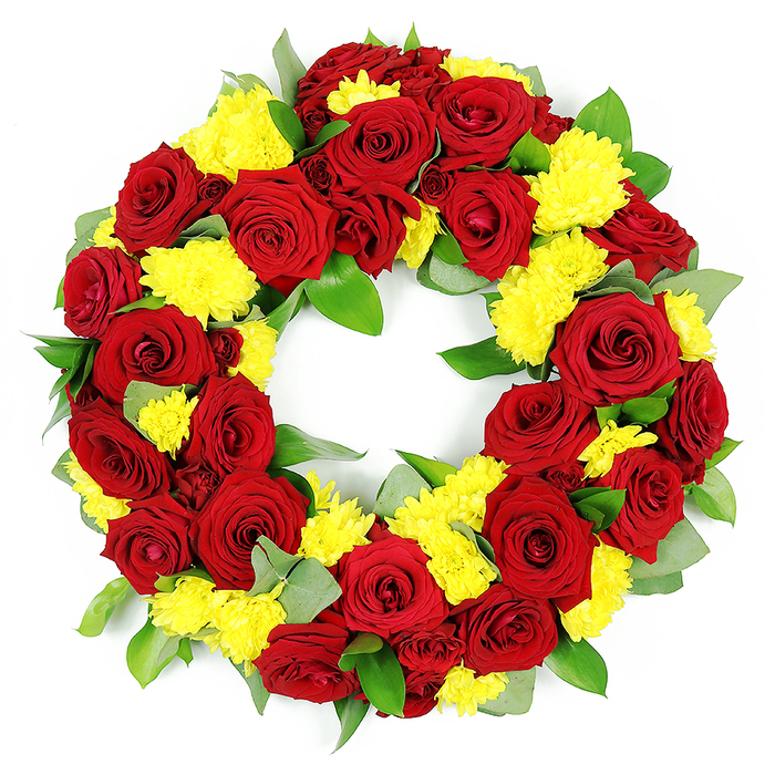 Against a bright white background, this circular floral wreath appears both bold and tender, its colours standing out with clarity while the form remains soft and rounded. The flowers are arranged densely in a neat ring, leaving a clear open centre that reinforces the traditional symbol of eternity and unbroken remembrance. Dominating the composition are deep red roses, carefully spaced around the wreath. Each bloom is fully open, revealing concentric layers of velvety petals that curl gently at the edges. The red tones are rich and complex, shifting from a classic crimson across the outer petals to darker, almost burgundy shades in the shadows, which gives the wreath a luxurious depth and a sense of solemn dignity. Between and around these roses are clusters of small yellow chrysanthemums, likely button or spray types, that create soft, cushion-like domes. Their many slender petals reach outwards in all directions, catching light and giving the yellow a lively, almost glowing quality. They introduce an uplifting note, suggesting warmth, gratitude, and cherished memories. A generous mix of foliage threads through the design, with robust dark-green leaves and softer, lightly frosted sprigs similar to eucalyptus providing structure and gentle movement. The greenery frames the flowers without hiding them, adding a natural balance and fullness. The even lighting makes the wreath appear freshly made, as though it could have just been completed in a Tolworth flower shop, ready to accompany a funeral cortege or a quiet farewell gathering. This arrangement feels well suited to local services in Tolworth, expressing sympathy, enduring love, and thoughtful remembrance through a simple yet striking combination of red roses and yellow chrysanthemums.