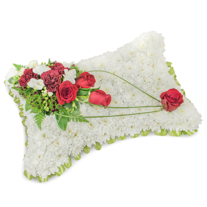 A poignant rectangular sympathy cushion, laid out on a neutral backdrop, whose face is entirely covered with hundreds of immaculate white chrysanthemums, packed tightly to create a lush, tactile surface that suggests calm and repose. The tiny, layered petals form a soft, wool-like texture that reads visually as both delicate and substantial. Anchoring the arrangement is an ornate spray at the top-left - several deep red roses in varied stages of bloom set against rich burgundy carnations - their velvety petals and dense ruffles forming a rich, warm counterpoint to the white base. Small white accent blooms, likely freesias or spray chrysanthemums, weave through the deep reds, adding bright, delicate points of light and a faint, honeyed scent that softens the composition. Verdant greenery, including slender ferns and broader glossy leaves, creates a supporting backdrop and helps the reds sing against the white. From the main cluster a trio of arcing green stems or bear-grass strands flows diagonally across the cushion toward the lower-right, where a single, prominent red rose rests as a quiet punctuation - an emblem of enduring love and remembrance. A subtle lime-green ribbon border peeks around the slightly raised edge, lending a neat, finished frame to the piece. This cushion reads as an understated, respectful memorial tribute familiar to families in Tolworth and neighbouring Kingston upon Thames, an arrangement chosen for funeral services, chapel displays, or graveside remembrance when one wants an expression of deep affection combined with serene simplicity.