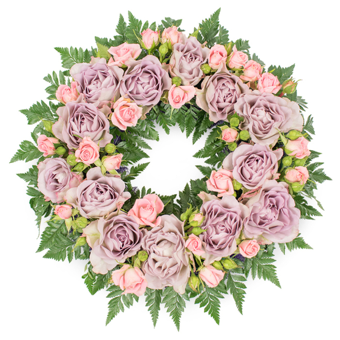 This image presents a beautifully structured funeral wreath, photographed straight from above on a pristine white surface so the composition appears perfectly circular and balanced. The foundation is a dense collar of dark green fern fronds, their serrated edges and finely divided leaflets overlapping in layers, giving the impression of depth, softness, and natural woodland texture. Rising from this green base, fully open roses in nuanced shades of dusty lavender, antique mauve, and soft, grey-tinted purple form the primary rhythm of the design. Each rose is lush and voluminous, its many petals swirling inward to a slightly shadowed center, conveying a sense of quiet richness. Between these larger roses, clusters of smaller blush-pink spray roses and delicate rosebuds are interwoven, some buds still round and closed, others beginning to open, revealing gentle inner petals. These pastel pink accents bring warmth and lightness to the cooler purples and help create a tapestry-like feel across the wreath. Subtle infill of darker greenery and tiny buds is tucked discreetly between the flowers, providing contrast and a more organic, garden-grown impression. The colour palette, with its interplay of soft purples, tender pinks, and steadfast greens, evokes feelings of sympathy, remembrance, and admiration, making it an appropriate tribute for a farewell gathering in Tolworth or along the nearby Kingston Road. The lighting is soft and evenly distributed, illuminating the petal textures and the glossy surfaces of the leaves without harsh glare. Overall, the wreath exudes thoughtful craftsmanship and emotional sensitivity, as though arranged by a local florist attuned to the needs of grieving families looking for a gentle, dignified floral expression.