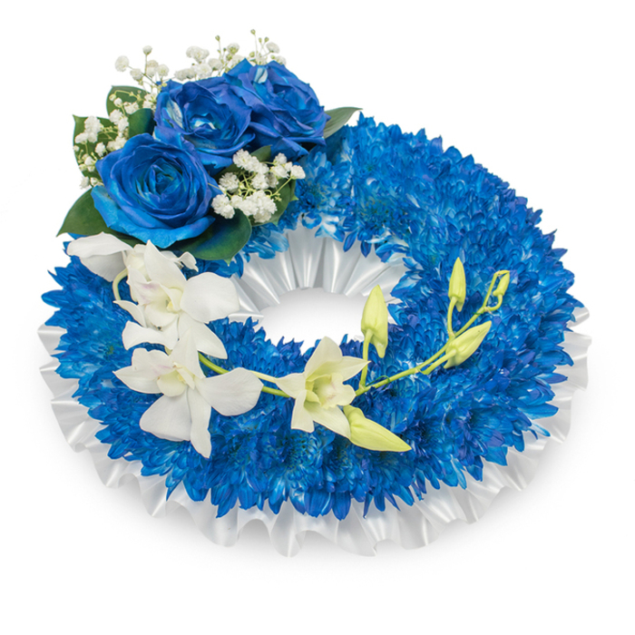 Framed against a pristine white background, this funeral wreath is a study in composed reverence, dominated by saturated blue and immaculate white. The circular form is densely filled with countless small, deep-blue chrysanthemum-like flowers, their fine, bristled petals overlapping to create a voluminous, tactile ring that feels both substantial and comforting. Lining the inner circumference is a pleated white satin ribbon-softly ruffled and glossy-which provides a refined backdrop and a subtle visual margin between the blue floral mass and the wreath's hollow centre. Prominently placed at the upper-left curve is a cluster of three large blue roses; their rich, sapphire-hued petals display a velvety surface and layered openness that contrasts beautifully with the chrysanthemums' compact texture. Accenting the roses are sprigs of tiny white baby's breath and a few deep green leaves, which add classic floral balance and a touch of garden-fresh depth. A gentle cascade descends from the mid-left, moving downwards and arcing toward the lower right: pristine white orchid blooms-likely Phalaenopsis-sit along a slender stem, their smooth, papery petals and delicate lips introducing an ethereal quality. Scattered among them are pale green orchid buds in various stages of opening, symbolising continuity and new growth amid grief. The lighting is soft and even, highlighting the interplay of matte and glossy surfaces without harsh shadows. Crafted by Flowers Tolworth for memorial services or condolence displays, this wreath communicates respectful solemnity, purity of sentiment, and a quiet promise of remembrance for local funerals and commemorations.
